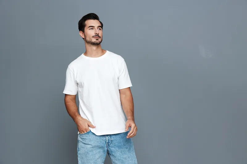 Stylish young man in casual white t-shirt and blue jeans standing against solid grey background, looking confidently into the distance. Fashion trend concept.