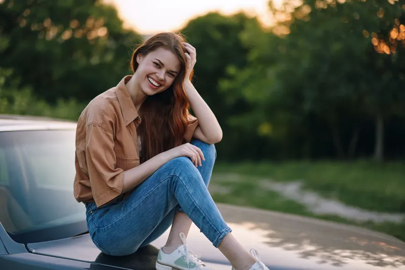 Smiling young woman with long hair sitting on a car hood, wearing casual outfit, enjoying a sunny day in a green outdoor setting.