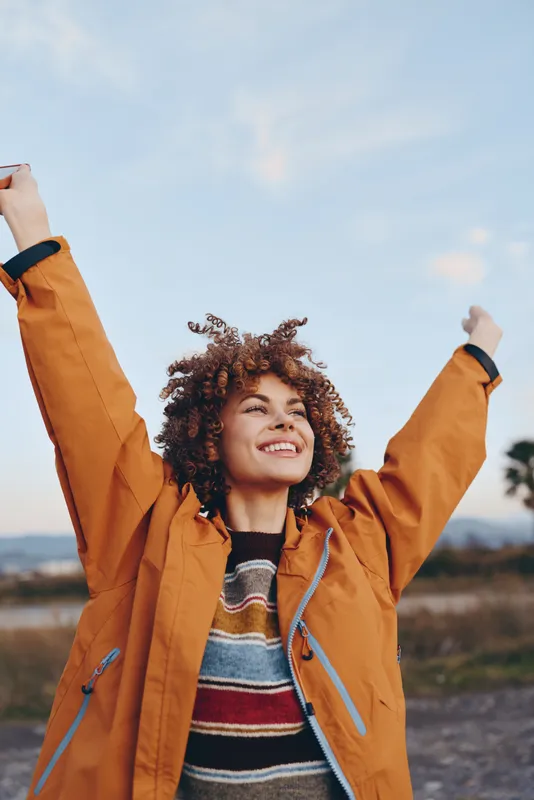 woman lifestyle outdoors, curly hair smile, arms raised, orange jacket on, enjoying fresh air in nature, scenic sky background, casual autumn joy, soft wind golden light today
