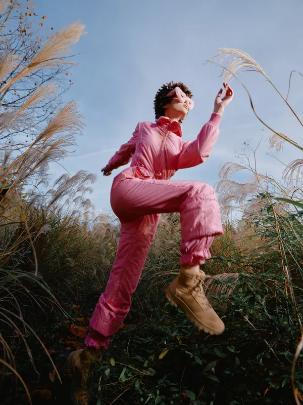 Woman jumping outdoors in a pink jumpsuit, boots and short hair, leaping through tall grass against a bright blue sky, conveying energy, playfulness, and free spirit in outdoor fashion.