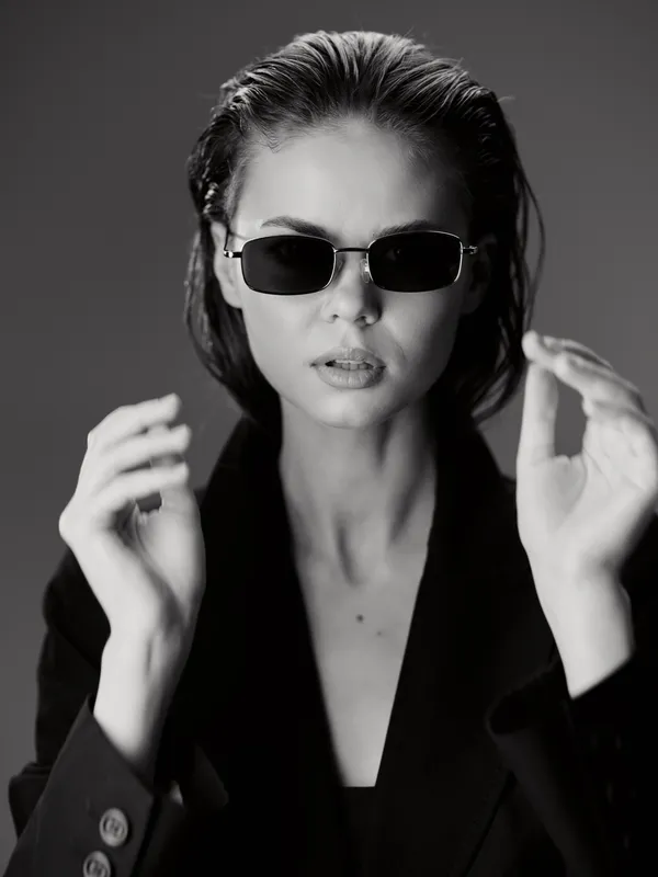 Black and white portrait of a stylish woman wearing sunglasses and a blazer in moody lighting.