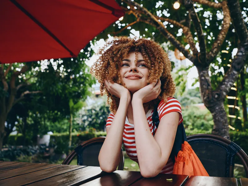 Young woman with curly hair sits at an outdoor cafe table under a red umbrella, smiling with hands cupping her face, wearing a striped shirt in a sunny garden.