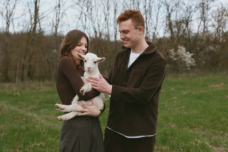 Joyful couple outdoors smiling while holding a young goat, tender interaction in a sunlit field, soft breeze, natural colors, candid lifestyle moment, warm countryside vibe, wholesome and charming