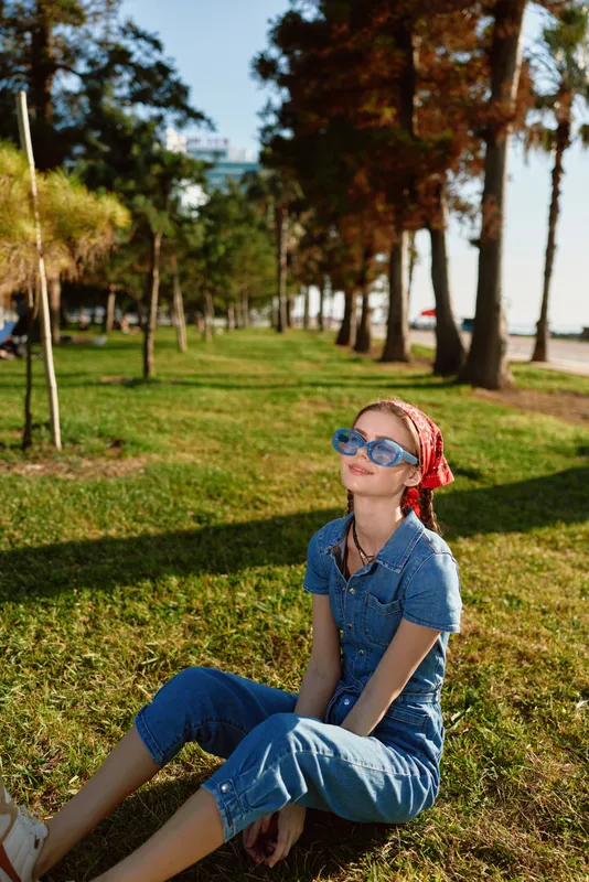 Lifestyle woman in modern boho-western denim street style sits on grass outdoors with sunglasses and red bandana, enjoying sunny day in film color tones.