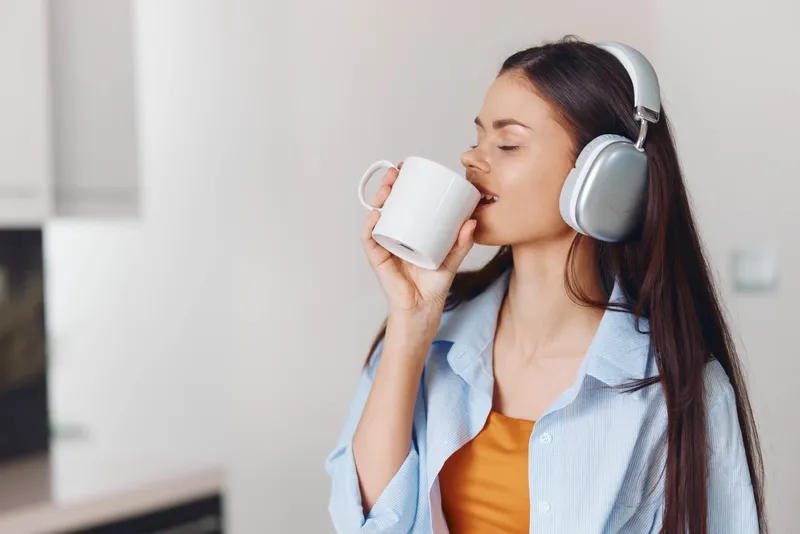 Женщина на кухне в наушниках пьет кофе и слушает музыку на телефоне
