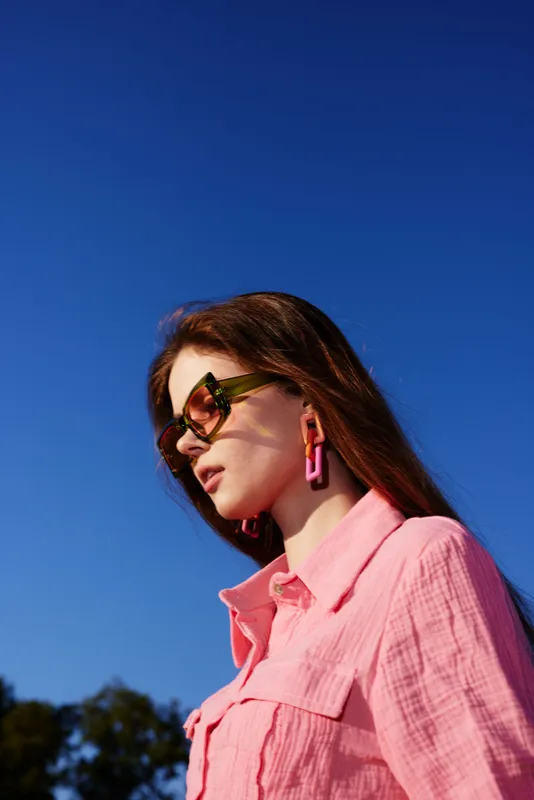 Stylish young woman in pink shirt and sunglasses against a clear blue sky, showcasing modern fashion and vibrant summer vibes
