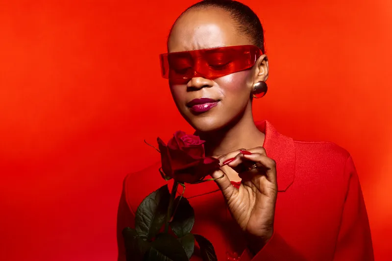 Portrait of elegant black woman with dark skin wearing red sunglasses and red outfit holding red rose against bright red background. Concept of fashion, beauty, and style.