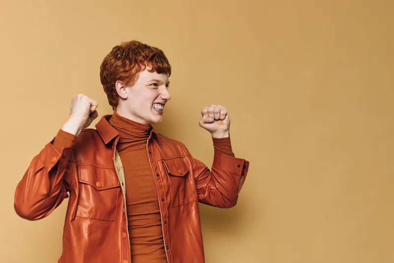 Young man with red hair wearing brown leather jacket celebrates success with clenched fists and joyful expression on beige background, showing excitement and confidence.