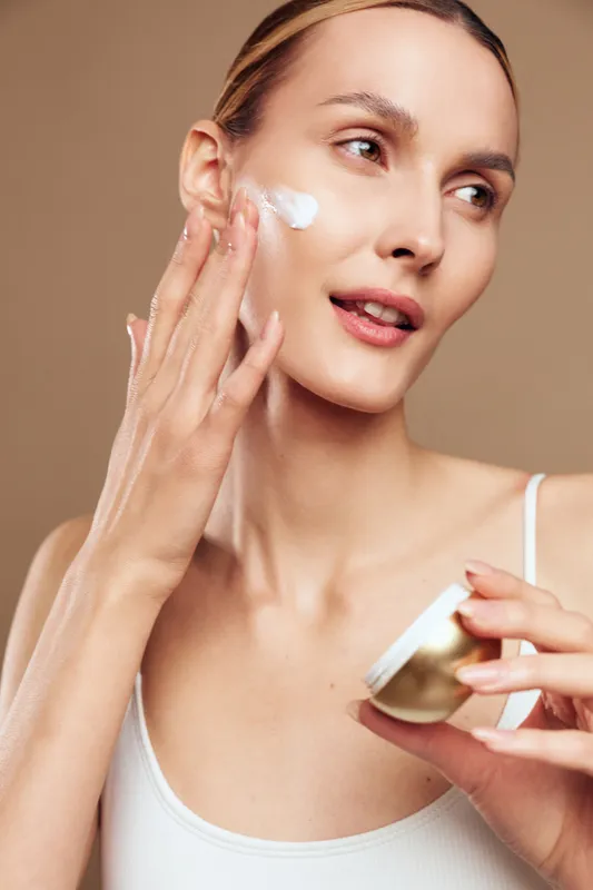 Woman applying white face cream with fingers, soft studio lighting, close portrait on brown background. Skincare selfcare and wellness concept, smooth skin care routine daily care.