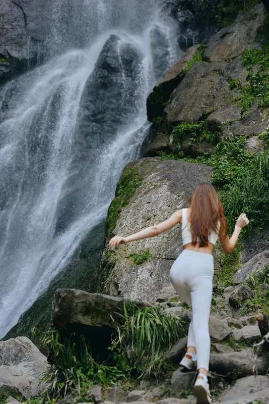 woman hiking near waterfall in athletic wear climbs rocky trail surrounded by greenery and flowing water in outdoor adventure scene
