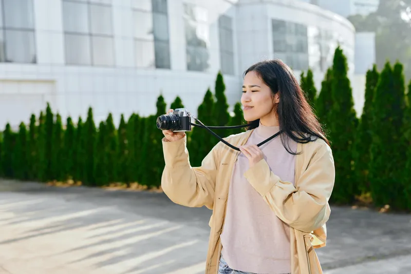 Woman holds camera on sunny outdoor street, casual jacket, evaluating shot with focused expression, urban backdrop, greenery, daylight, candid photography moment