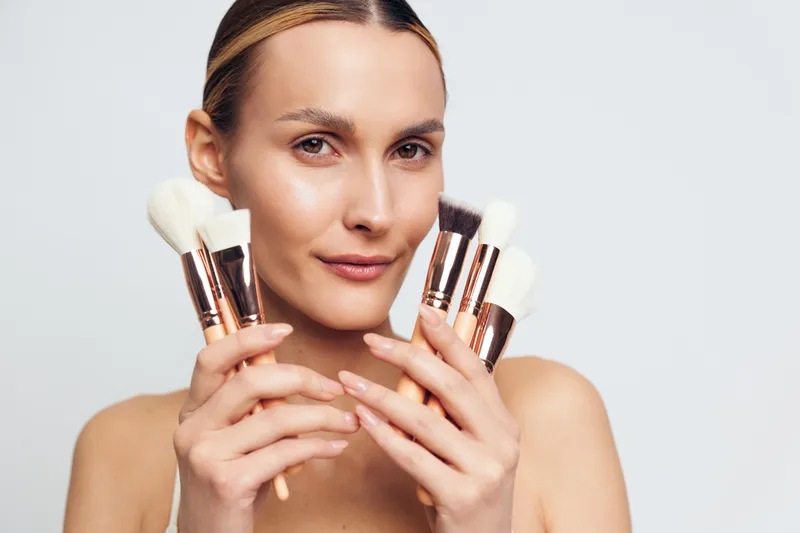 Young woman holding various makeup brushes close to face, neutral expression, clean skin, studio lighting, white background, beauty tools, cosmetic brushes.