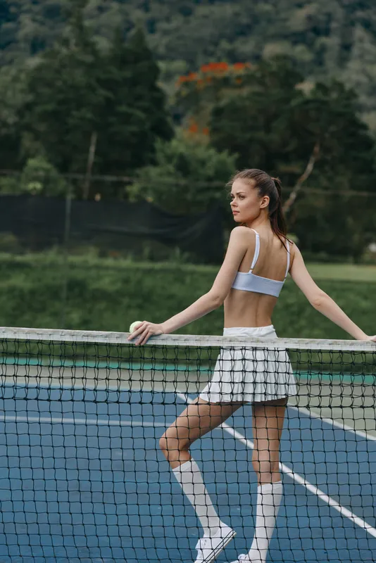 Young female tennis player in sporty attire striking a confident pose by the net, showcasing a determined expression in a vibrant outdoor setting