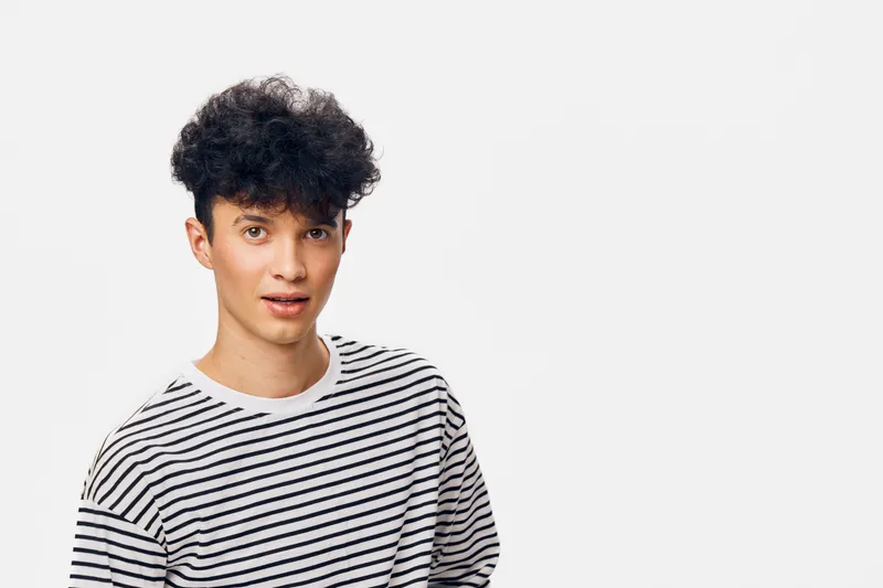 Young man with curly hair in striped shirt looking surprised against white background, casual style, natural light, studio portrait.