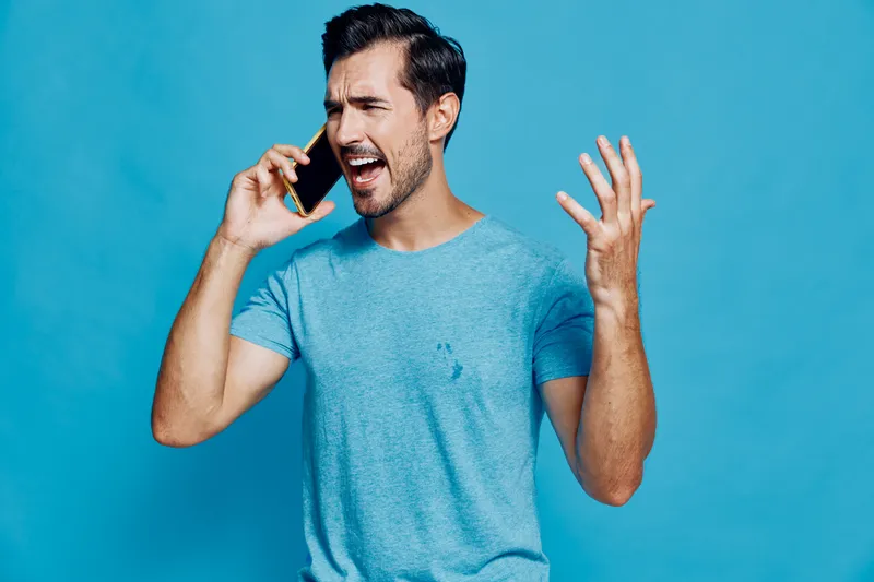 Expressive man talking on mobile phone, looking frustrated with hands raised, wearing casual light blue t-shirt, set against a vibrant blue background.