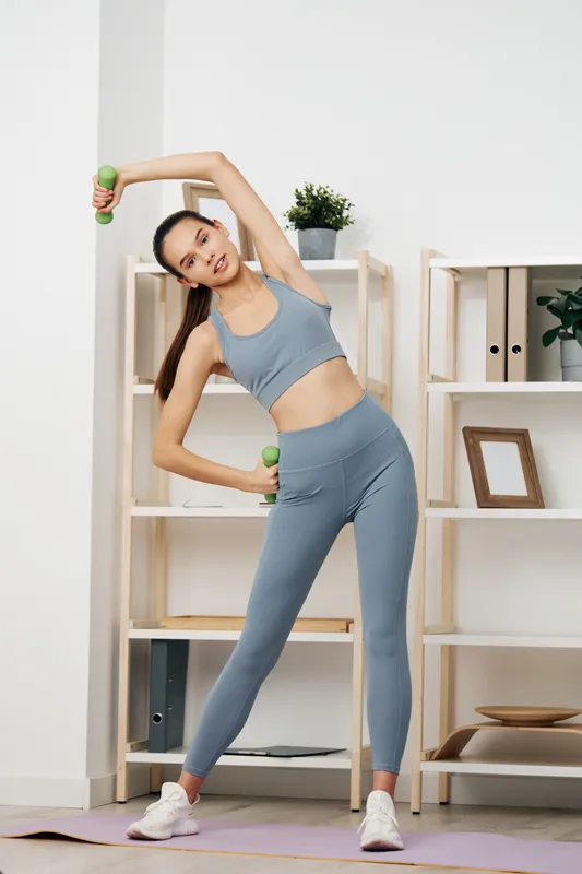 Woman in blue athletic wear performing side stretch with dumbbells in a bright home gym, shelves and plants in background, showcasing fitness, balance and focused exercise.
