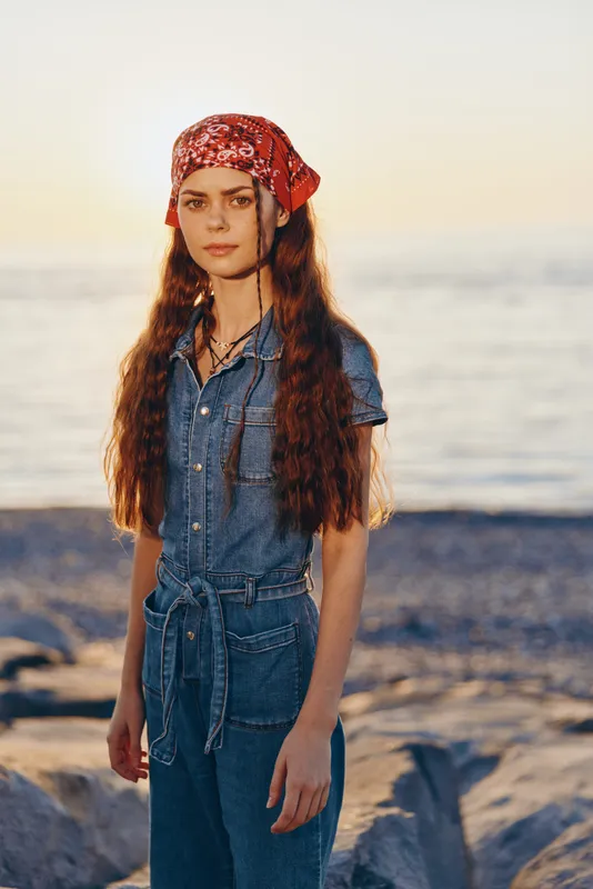 young woman with long hair wearing denim jumpsuit and red bandana standing on rocky beach at sunset with calm sea in background