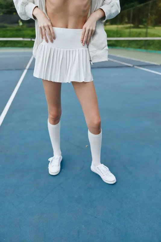 Young athletic female model in a white tennis outfit, showcasing playful emotions at a tennis court in a summer setting, surrounded by green nature