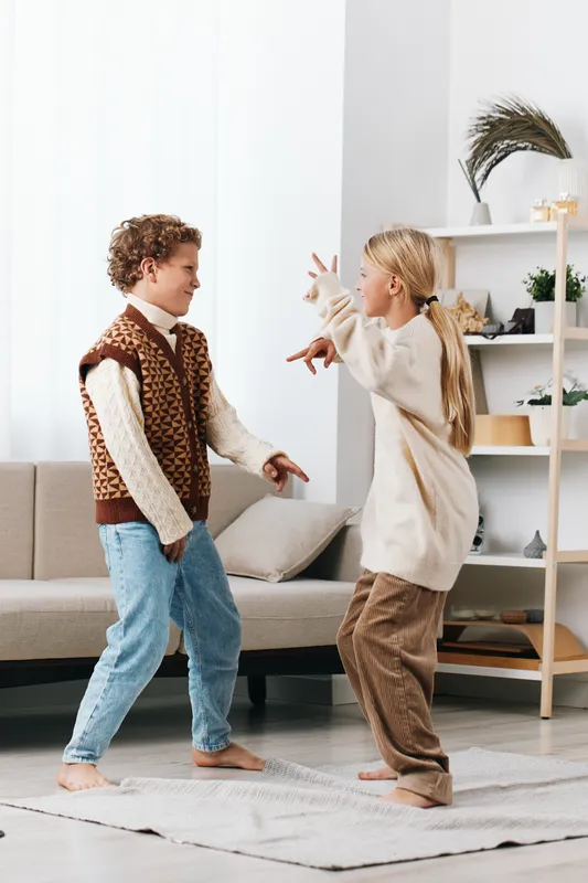 Two children playfully dance in a bright living room, wearing cozy sweaters and jeans, mid motion with expressive gestures and smiles, capturing candid, joyful movement.