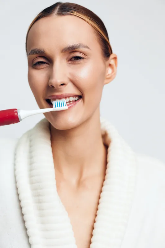 Smiling woman brushing her teeth with a red toothbrush, wearing a white robe, close up on a light background, concept of oral care, hygiene, and daily routine, bright studio lighting