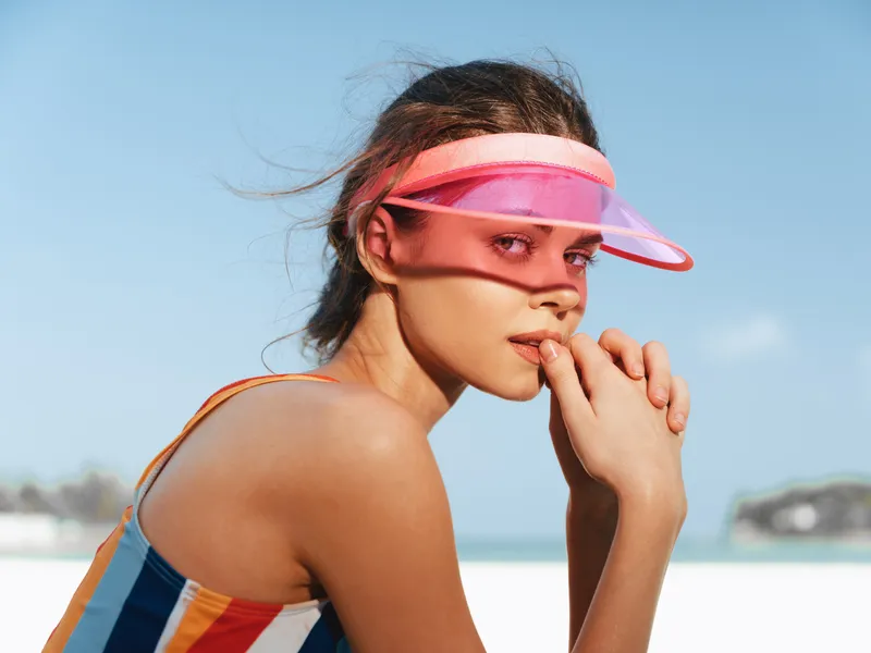 A stylish woman wears a pink visor at a sunny beach, looking toward the distance with a thoughtful expression while resting her chin on her hand, relaxed and poised.