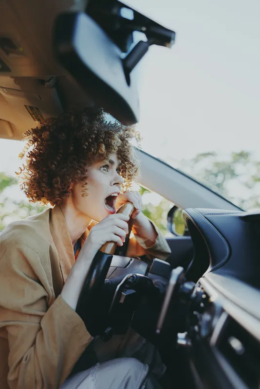 Curly haired woman eating a snack inside a car, daytime natural light highlights warm retro tones, casual lifestyle mood, candid moment during a daytime drive, soft, warm glow.