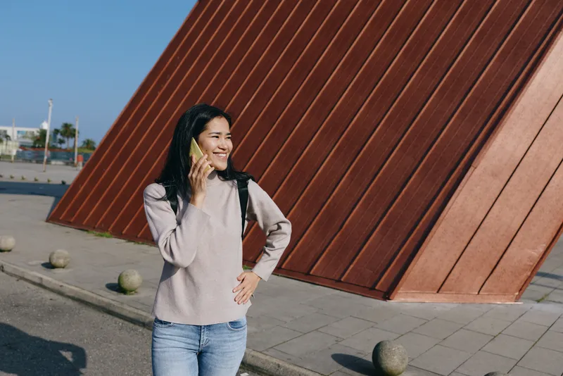Woman with phone in hand, outdoors beside a bold angular rust wall, wearing a light sweater and jeans, smiling while chatting in a sunny, modern street setting.