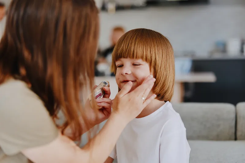 Loving woman applying skincare to her son’s face, ensuring he has healthy skin and a joyful smile. A warm and caring moment that captures family bonding.