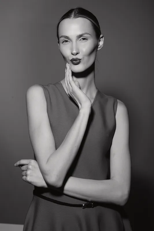 Slim Caucasian woman with pale skin and slicked back hair poses with hand on face in black and white studio portrait.