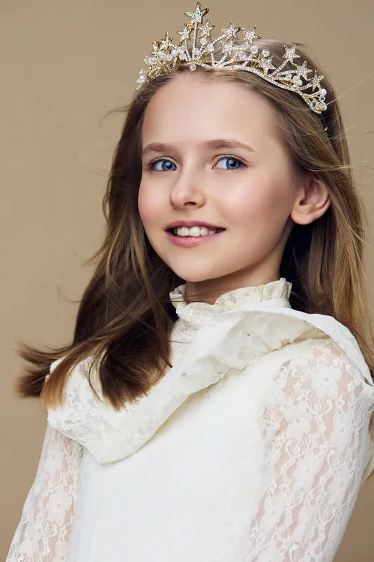 Young girl with a crown, wearing a stylish lace dress, smiling joyfully against a soft beige background, highlighting childhood elegance and innocence