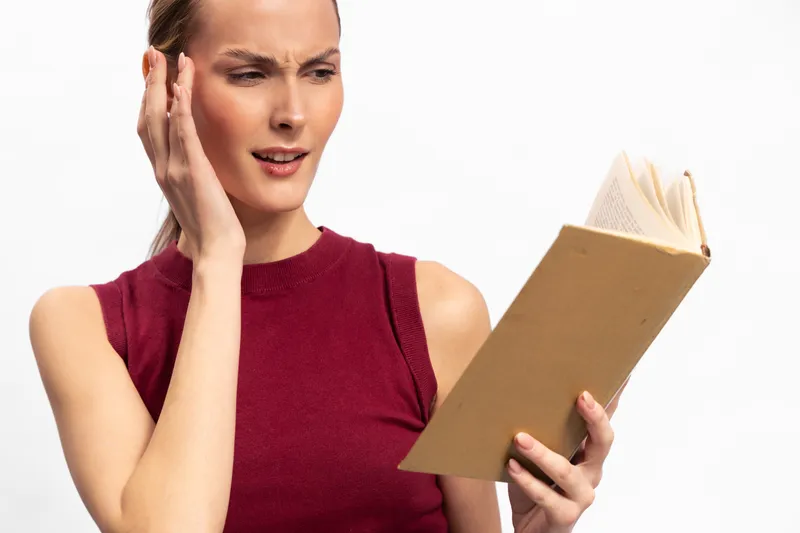 Confused young woman in red sleeveless top holds open book and touches ear, isolated on white. Concept of difficulty reading, misunderstanding, and concentration problems.