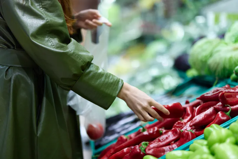 Женщина в продуктовом магазине выбирает свежие овощи для приготовления ужина дома, держа в руках красный перец во время покупок на рынке