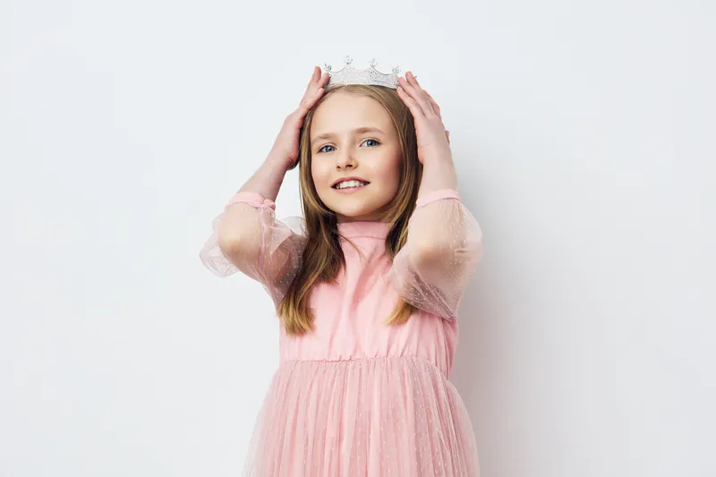 Happy young girl in a pink dress and crown, celebrating childhood joy with a cheerful expression against a light background Perfect for children related themes