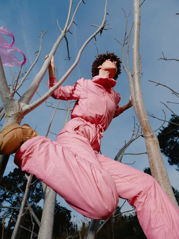 Outdoor scene with a person in a pink jumpsuit climbing among bare trees against a clear blue sky, capturing bold aerial pose and dynamic movement in nature.
