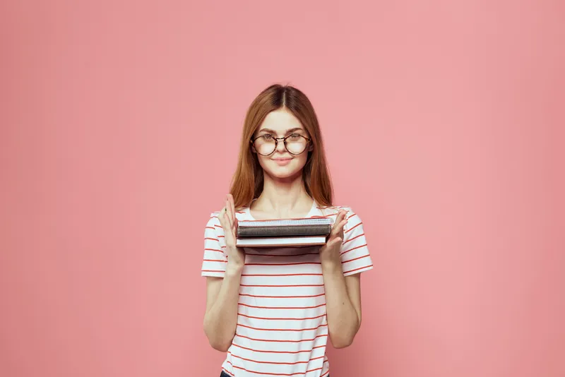 Молодая студентка с книгами на розовом фоне, в очках, крупный план лица