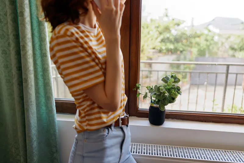 A young woman in a striped orange top stands by a sunlit window, touching her hair and relaxing with a calm, thoughtful expression while the plant on the sill adds a natural touch.