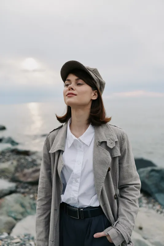 Young woman in casual coat and cap stands calmly on rocky shore near sea, eyes closed, enjoying peaceful breeze. Concept of relaxation, nature, and outdoor lifestyle.