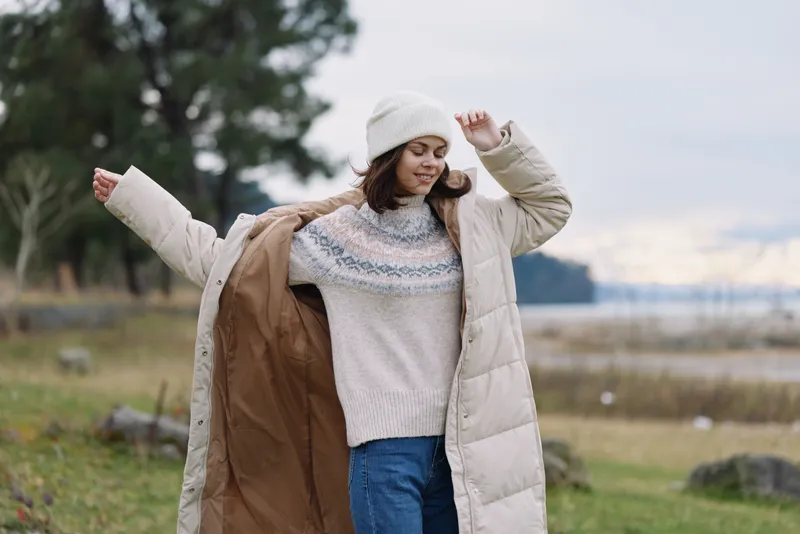 Woman wearing a long beige puffer coat and knit sweater outdoors, enjoying a windy day by the shore, casual winter fashion and cozy cold weather style for a relaxed lifestyle shoot