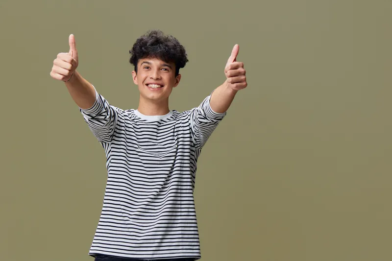 smiling young man wearing striped shirt showing thumbs up gesture with both hands against neutral background expressing positivity and confidence