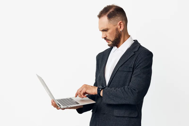A focused business professional man stands against an isolated colored background, using a laptop with calm confidence and focused expression.