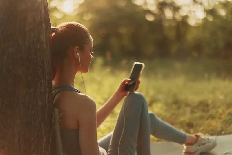 Young woman listening to music outdoors in casual athletic wear, enjoying a sunny afternoon while using her smartphone, representing a healthy lifestyle and leisure