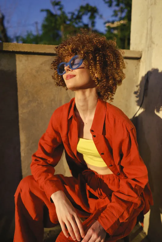 Cheerful woman outdoors, wearing bold orange outfit, curly hair, sunglasses, sunlit moment, relaxed pose, warm tones, natural setting against warm wall backdrop