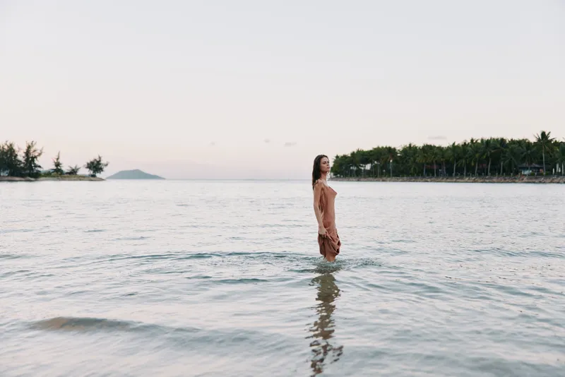 A solitary figure stands waist-deep in tranquil shoreline water, bathed in soft pastel light. The calm sea, gentle ripples, and open horizon create a serene, contemplative moment.