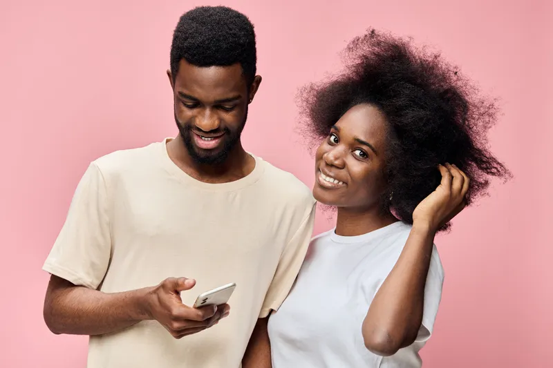 A joyful couple with natural hairstyles smiles together against a pink background, showcasing diversity, connection, and modern lifestyle with a phone