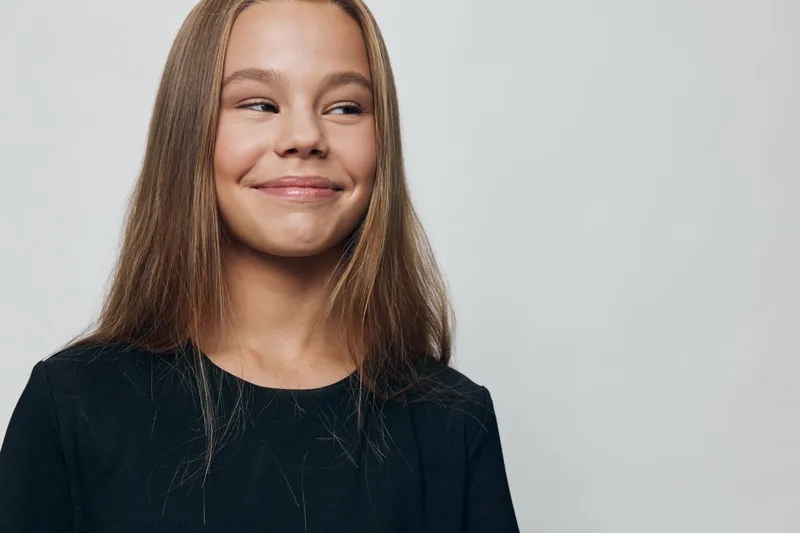 Girl with long hair and black shirt smiling and looking sideways on plain background for positive emotion and happiness with casual style and natural light.