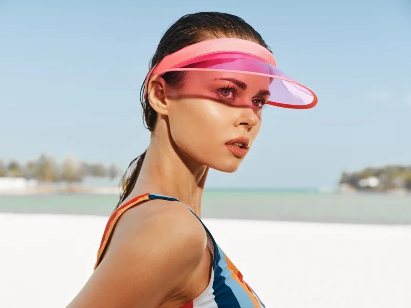 Portrait of a stylish model on a sunny beach wearing a pink visor and colorful swimsuit, capturing a crisp, fashion-forward moment against a bright horizon.