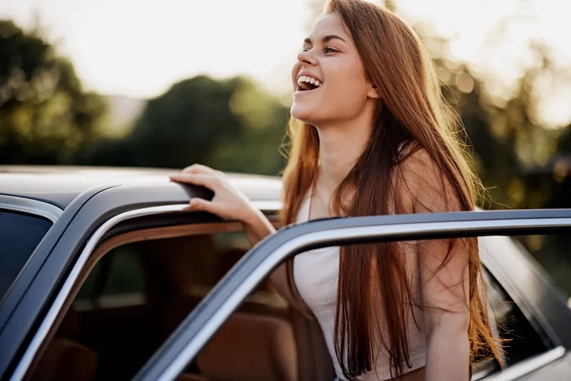 Happy young woman with long brown hair enjoying a sunny day by the car, wearing a casual outfit and smiling in a vibrant outdoor setting.