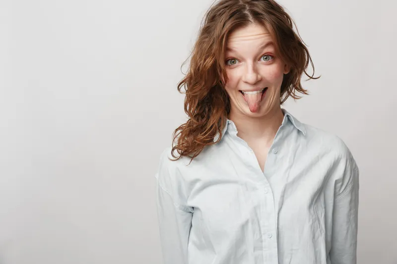 Young woman with curly hair wearing white shirt sticks out tongue playfully. Studio portrait on grey background. Concept of fun, humor, youth, and casual lifestyle expression.
