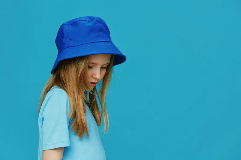A young girl wearing a blue bucket hat and a matching t shirt, looking down against a solid blue background, embodying a calm and thoughtful mood