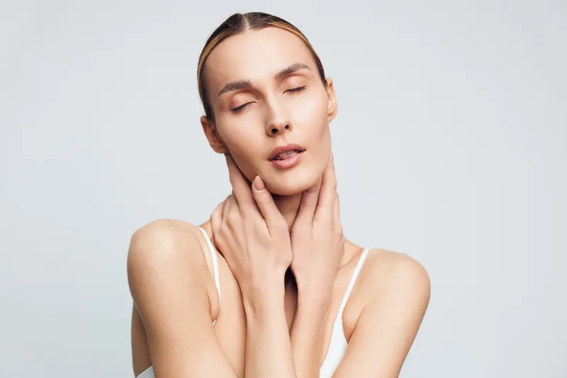 Young woman with closed eyes gently touches her neck and face while standing against a plain light background in a soft white tank top, conveying calmness and self-care.